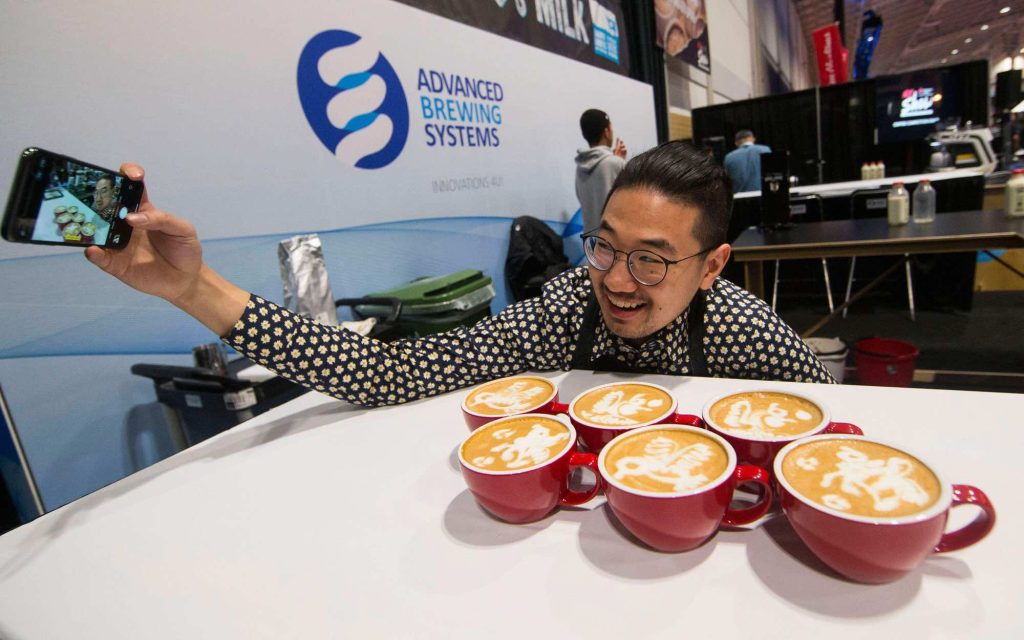 Pessoa fazendo selfie junto a xícaras de cappuccino com latte art.