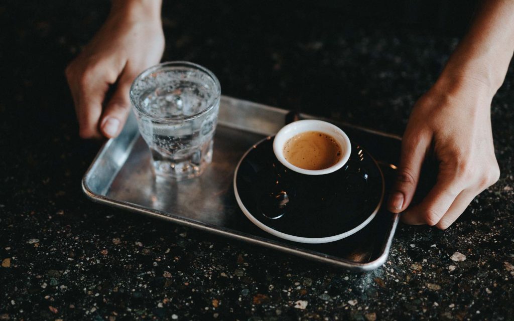 A barista holds a tray with a glass of water and an espresso.