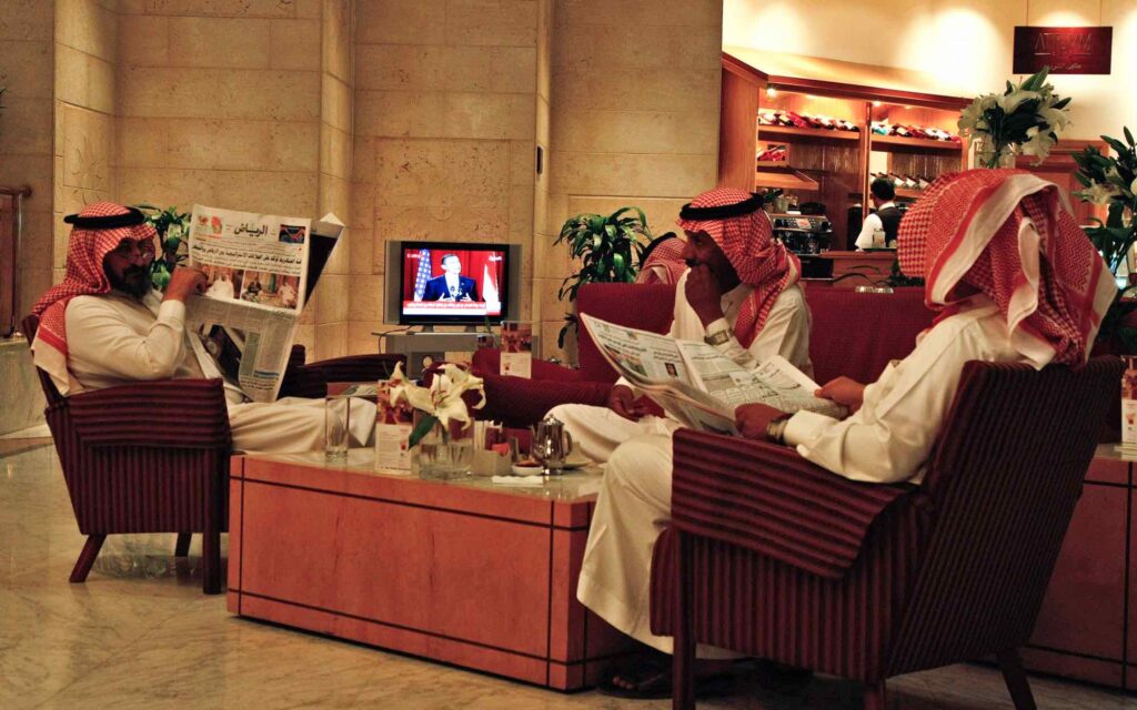 Men read newspapers and drink beverages in a hotel lobby.